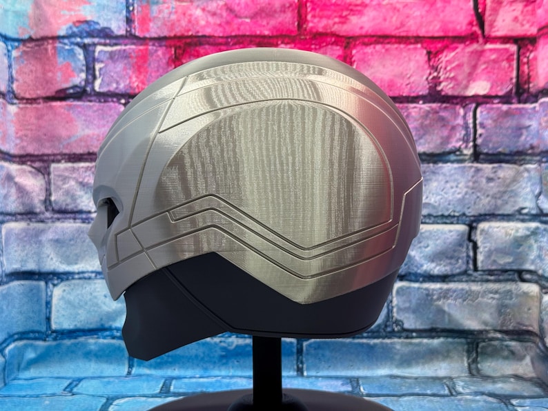May include: A silver and gray helmet with a textured metallic finish. The helmet has a streamlined design with angular details and a dark gray lower section. It is displayed on a black stand against a brick wall.
