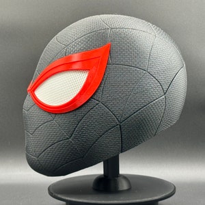 Puede incluir: Una m&aacute;scara de Spider-Man impresa en 3D de color negro y rojo con una pieza de ojo de malla blanca. La m&aacute;scara est&aacute; en un soporte negro.