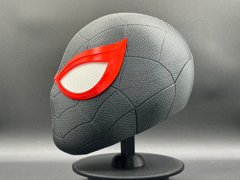 Puede incluir: Una m&aacute;scara de Spider-Man impresa en 3D de color negro y rojo con una pieza de ojo de malla blanca. La m&aacute;scara est&aacute; en un soporte negro.