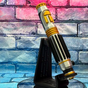 May include: A silver and gold lightsaber prop with black accents, displayed on a black stand. The lightsaber features a cylindrical design with ribbed sections and yellow buttons. The background is a brick wall with pink and blue hues.