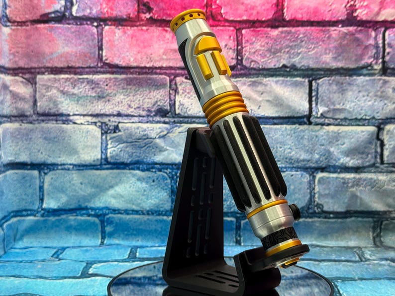 May include: A silver and gold lightsaber prop with black accents, displayed on a black stand. The lightsaber features a cylindrical design with ribbed sections and yellow buttons. The background is a brick wall with pink and blue hues.