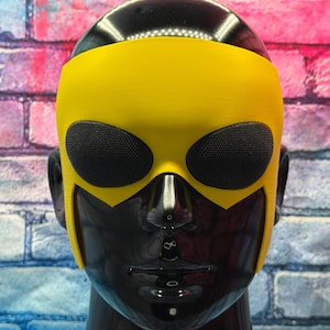May include: A bright yellow mask featuring two oval-shaped black mesh eye coverings. The mask is fitted onto a glossy black mannequin head, covering the upper portion of the face and extending down the sides to frame the cheekbones and ears.