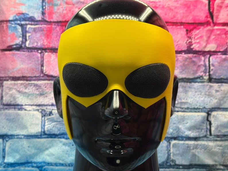 May include: A bright yellow mask featuring two oval-shaped black mesh eye coverings. The mask is fitted onto a glossy black mannequin head, covering the upper portion of the face and extending down the sides to frame the cheekbones and ears.
