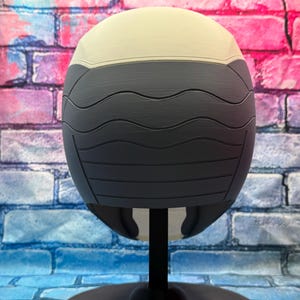 May include: A two-tone, spherical object with a cream-colored top and a dark gray, textured lower section. The gray portion features horizontal, wave-like grooves. The object is mounted on a black stand, suggesting a display or decorative item.