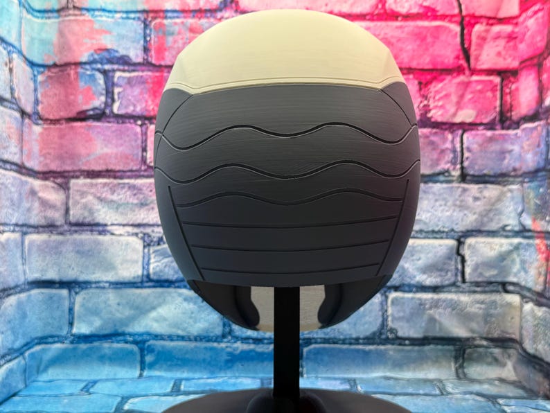 May include: A two-tone, spherical object with a cream-colored top and a dark gray, textured lower section. The gray portion features horizontal, wave-like grooves. The object is mounted on a black stand, suggesting a display or decorative item.