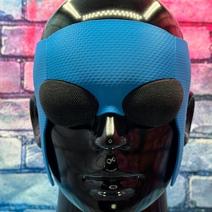 May include: A bright blue textured headband with two large, dark, oval-shaped mesh eye coverings positioned over the eyes of a glossy black mannequin head. The headband features a fine, repeating geometric pattern across its surface.