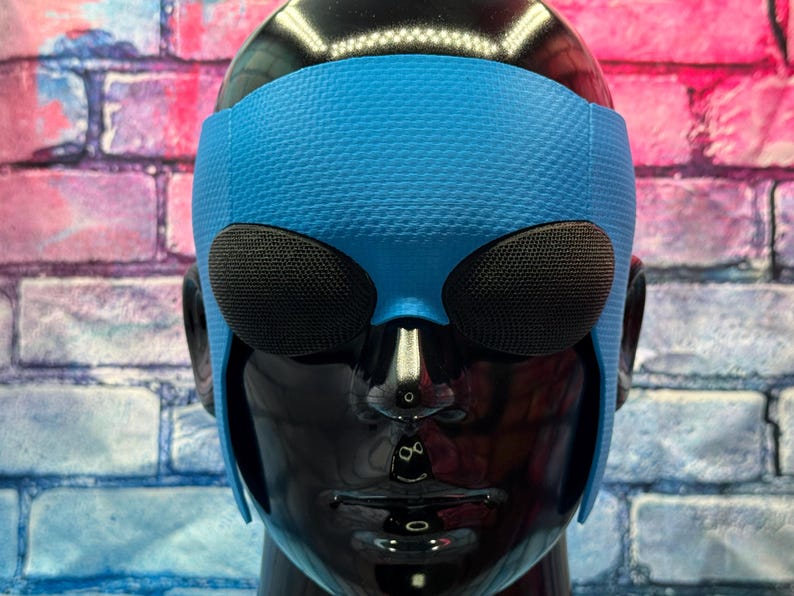 May include: A bright blue textured headband with two large, dark, oval-shaped mesh eye coverings positioned over the eyes of a glossy black mannequin head. The headband features a fine, repeating geometric pattern across its surface.