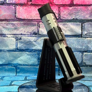 May include: A silver and black lightsaber prop, displayed on a black stand. The lightsaber has a cylindrical design with detailed panels and a black handle. The stand is angled, providing a clear view of the prop against a brick wall.