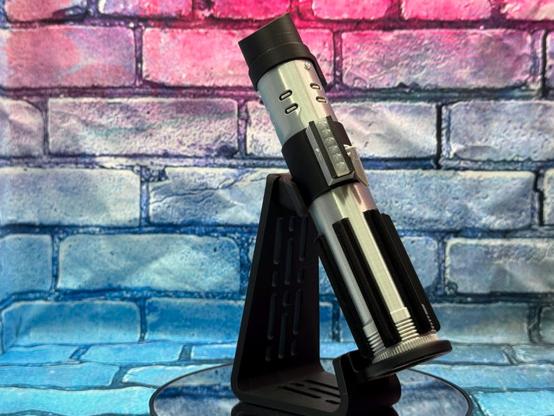 May include: A silver and black lightsaber prop, displayed on a black stand. The lightsaber has a cylindrical design with detailed panels and a black handle. The stand is angled, providing a clear view of the prop against a brick wall.