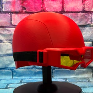 May include: A bright red helmet featuring a smooth, rounded dome shape and an integrated visor attachment. The visor is also red with yellow-tinted lenses and is secured to the helmet by a black elastic strap that wraps around the sides.