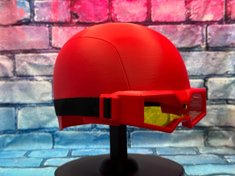 May include: A bright red helmet featuring a smooth, rounded dome shape and an integrated visor attachment. The visor is also red with yellow-tinted lenses and is secured to the helmet by a black elastic strap that wraps around the sides.