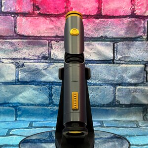 May include: A futuristic-looking, gray and gold device, possibly a lightsaber or a similar prop, is displayed against a brick wall backdrop. The device has a cylindrical body with gold accents and a button. It is mounted on a black stand.