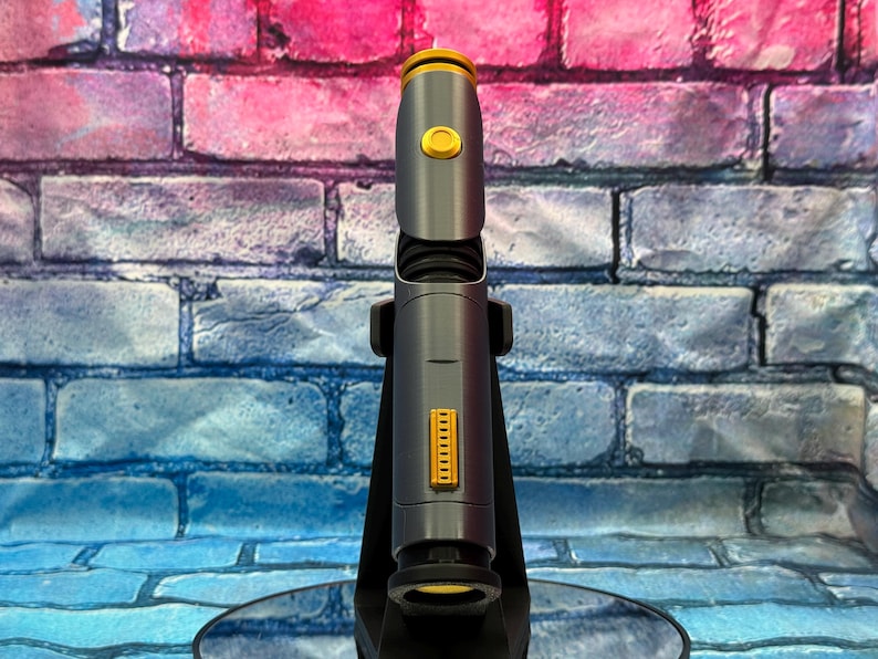May include: A futuristic-looking, gray and gold device, possibly a lightsaber or a similar prop, is displayed against a brick wall backdrop. The device has a cylindrical body with gold accents and a button. It is mounted on a black stand.