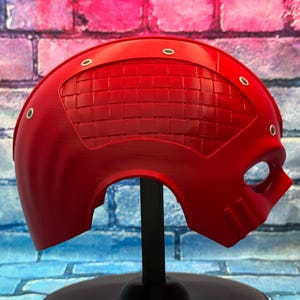 May include: A vibrant red helmet with a textured visor and eye opening, mounted on a black stand. The helmet has a smooth finish with several small, silver-colored rivets. The background is a colorful brick wall.