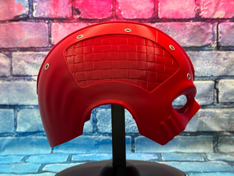 May include: A vibrant red helmet with a textured visor and eye opening, mounted on a black stand. The helmet has a smooth finish with several small, silver-colored rivets. The background is a colorful brick wall.