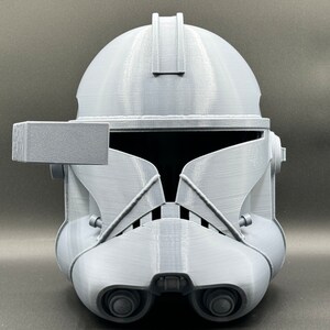 Puede incluir: Un casco gris de pl&aacute;stico impreso en 3D con una visera y un peque&ntilde;o accesorio rectangular en el lado izquierdo. El casco tiene una superficie texturizada y un dise&ntilde;o que se asemeja a un casco de soldado clon de Star Wars.