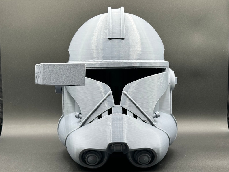 Puede incluir: Un casco gris de pl&aacute;stico impreso en 3D con una visera y un peque&ntilde;o accesorio rectangular en el lado izquierdo. El casco tiene una superficie texturizada y un dise&ntilde;o que se asemeja a un casco de soldado clon de Star Wars.