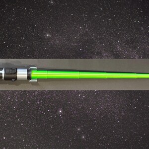 May include: A green lightsaber prop with a black and silver handle. The lightsaber is a toy or collectible item, likely from a science fiction franchise. The background is a starry night sky.