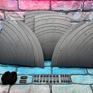 May include: A set of gray, curved, textured panels with horizontal lines, possibly for a diorama or display. Also included are black straps and small rectangular pieces. The items are arranged on a blue and gray brick surface, with a brick wall backdrop.