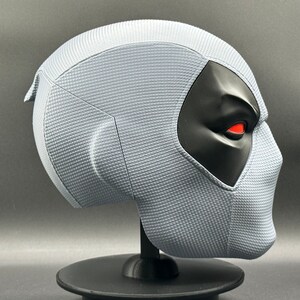 Może przedstawiać: Biała maska Deadpoola z czarną maską na oczy i czerwonymi oczami. Maska stoi na czarnym stojaku.