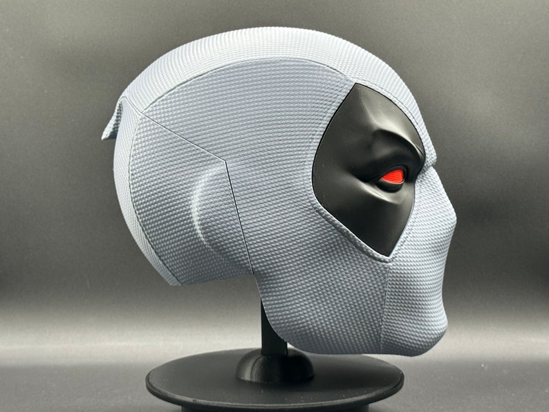 Może przedstawiać: Biała maska Deadpoola z czarną maską na oczy i czerwonymi oczami. Maska stoi na czarnym stojaku.