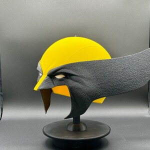 Puede incluir: Una m&aacute;scara de superh&eacute;roe impresa en 3D de color amarillo y negro. La m&aacute;scara est&aacute; en un soporte negro y tiene una superficie texturizada.
