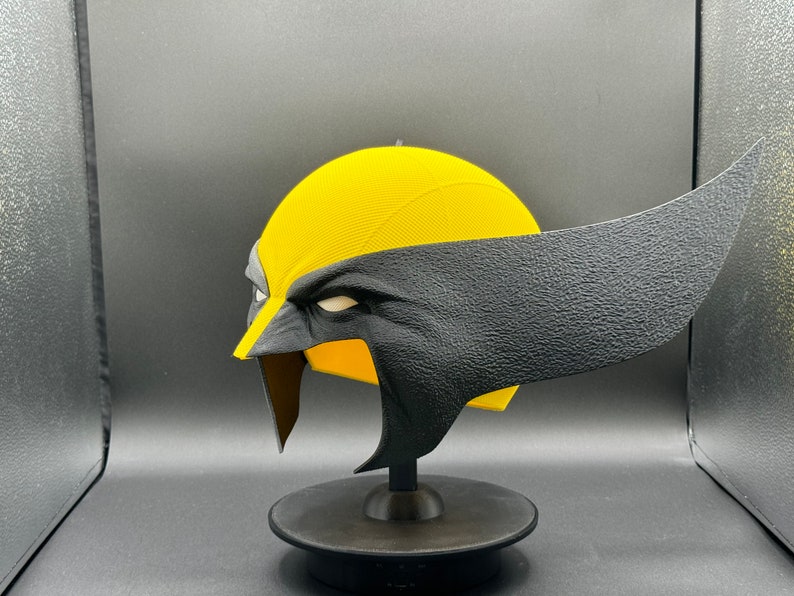 Puede incluir: Una m&aacute;scara de superh&eacute;roe impresa en 3D de color amarillo y negro. La m&aacute;scara est&aacute; en un soporte negro y tiene una superficie texturizada.