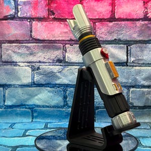 May include: A silver and black lightsaber prop with orange and red accents, displayed on a black stand. The prop has a detailed design with a ribbed grip and a blade emitter. The background is a brick wall with blue and pink hues.