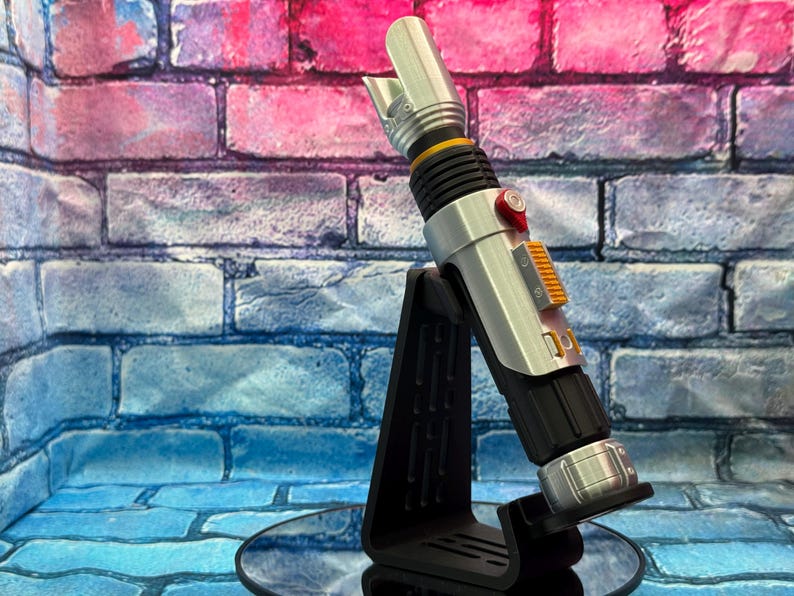 May include: A silver and black lightsaber prop with orange and red accents, displayed on a black stand. The prop has a detailed design with a ribbed grip and a blade emitter. The background is a brick wall with blue and pink hues.