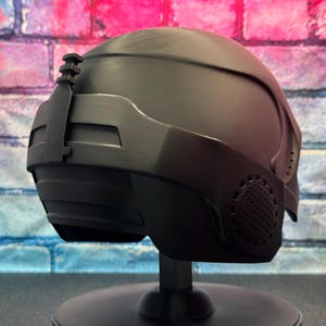 May include: A matte black helmet with a futuristic design, featuring a visor and side details. The helmet is displayed on a black stand against a brick wall background with pink and blue hues. The helmet has a textured surface and a streamlined shape.