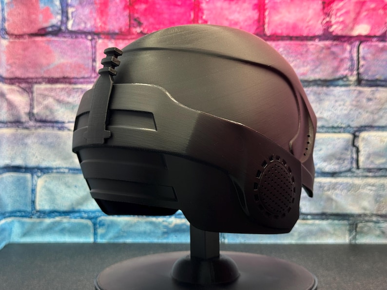 May include: A matte black helmet with a futuristic design, featuring a visor and side details. The helmet is displayed on a black stand against a brick wall background with pink and blue hues. The helmet has a textured surface and a streamlined shape.