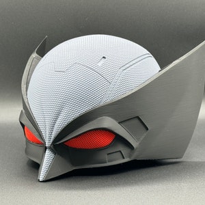May include: A black and gray 3D printed mask with red mesh eyes. The mask has a textured surface and a pointed chin.