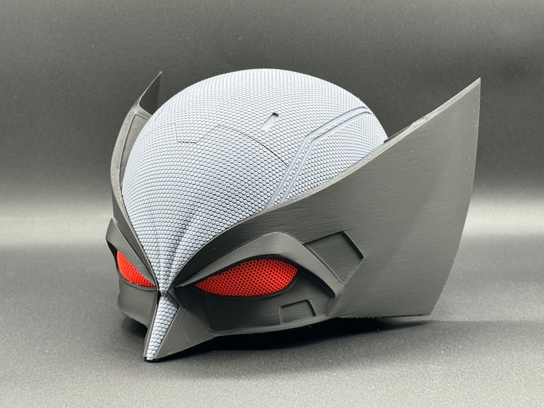 May include: A black and gray 3D printed mask with red mesh eyes. The mask has a textured surface and a pointed chin.