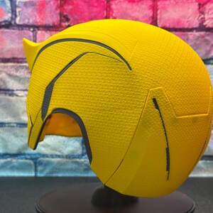 Puede incluir: Un casco amarillo brillante con detalles negros y una superficie texturizada. El casco tiene una parte superior puntiaguda y una visera curva. Se muestra sobre un soporte negro contra un fondo de pared de ladrillo.