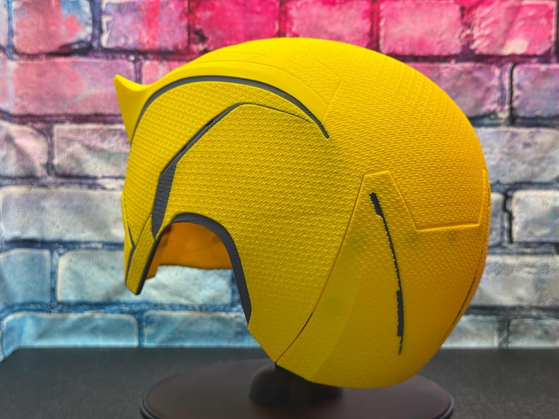 Puede incluir: Un casco amarillo brillante con detalles negros y una superficie texturizada. El casco tiene una parte superior puntiaguda y una visera curva. Se muestra sobre un soporte negro contra un fondo de pared de ladrillo.