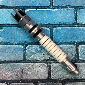 May include: A silver and white lightsaber prop with black accents. The hilt is white with black lines, and the top features a gray and black design. The prop is set against a blue brick wall.