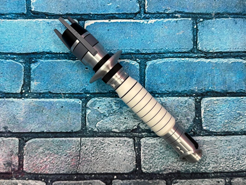 May include: A silver and white lightsaber prop with black accents. The hilt is white with black lines, and the top features a gray and black design. The prop is set against a blue brick wall.