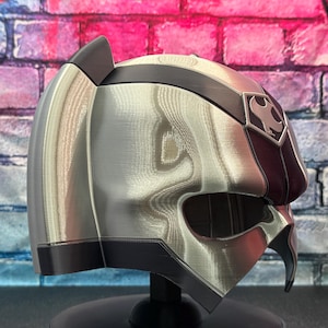 May include: A silver and black helmet with a unique design, featuring a dark red accent on the side and a symbol on the forehead. The helmet is displayed on a black stand, showcasing its intricate details and futuristic aesthetic.