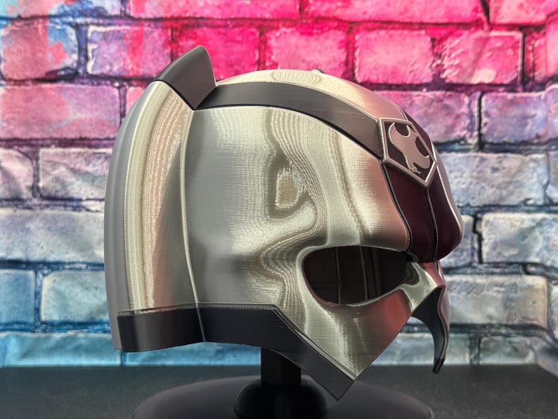 May include: A silver and black helmet with a unique design, featuring a dark red accent on the side and a symbol on the forehead. The helmet is displayed on a black stand, showcasing its intricate details and futuristic aesthetic.