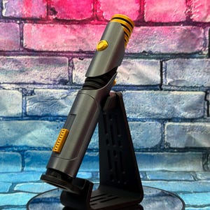 May include: A gray and yellow lightsaber prop, displayed on a black stand. The lightsaber has a cylindrical body with yellow accents on the hilt and emitter. The stand is angled, providing a clear view of the prop against a brick wall.