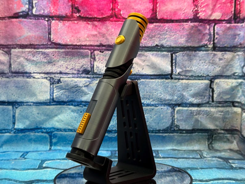 May include: A gray and yellow lightsaber prop, displayed on a black stand. The lightsaber has a cylindrical body with yellow accents on the hilt and emitter. The stand is angled, providing a clear view of the prop against a brick wall.