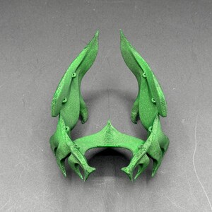 Puede incluir: Una escultura abstracta verde con un dise&ntilde;o sim&eacute;trico. La pieza presenta dos grandes formas en forma de hoja que se curvan hacia arriba, conectadas por un elemento central angular. Se aprecian peque&ntilde;os detalles circulares.