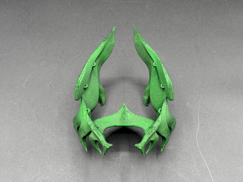 Puede incluir: Una escultura abstracta verde con un dise&ntilde;o sim&eacute;trico. La pieza presenta dos grandes formas en forma de hoja que se curvan hacia arriba, conectadas por un elemento central angular. Se aprecian peque&ntilde;os detalles circulares.