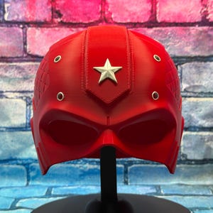 May include: A vibrant red helmet with a silver star emblem and circular accents. The helmet is displayed on a black stand against a brick wall background. The design suggests a superhero or cosplay theme.
