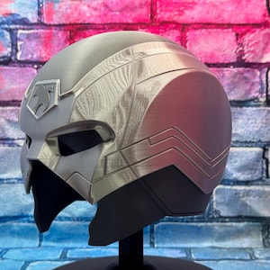 May include: A silver and gray helmet with a stylized design, featuring a prominent emblem on the forehead. The helmet has a sleek, futuristic aesthetic, with a dark visor area and a black base. The design includes textured panels and angular lines.