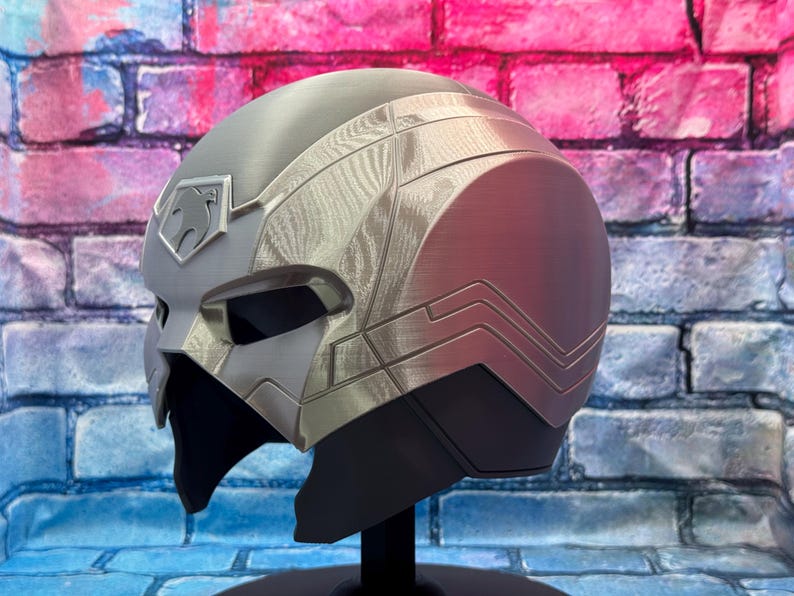 May include: A silver and gray helmet with a stylized design, featuring a prominent emblem on the forehead. The helmet has a sleek, futuristic aesthetic, with a dark visor area and a black base. The design includes textured panels and angular lines.