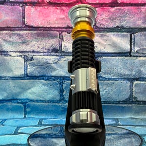 May include: A lightsaber prop in black, silver, and gold. The hilt has a ribbed design, silver details, and a gold section. It's displayed on a black stand against a brick wall. The background has blue, pink, and purple tones.