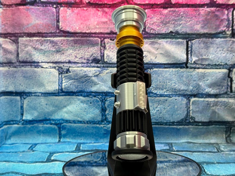 May include: A lightsaber prop in black, silver, and gold. The hilt has a ribbed design, silver details, and a gold section. It's displayed on a black stand against a brick wall. The background has blue, pink, and purple tones.