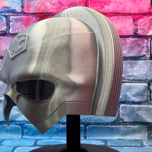May include: A silver-colored helmet with a Superman logo on the side. The helmet has a smooth, metallic finish and a distinctive design, including eye openings and a pointed chin. The helmet is displayed on a black stand.