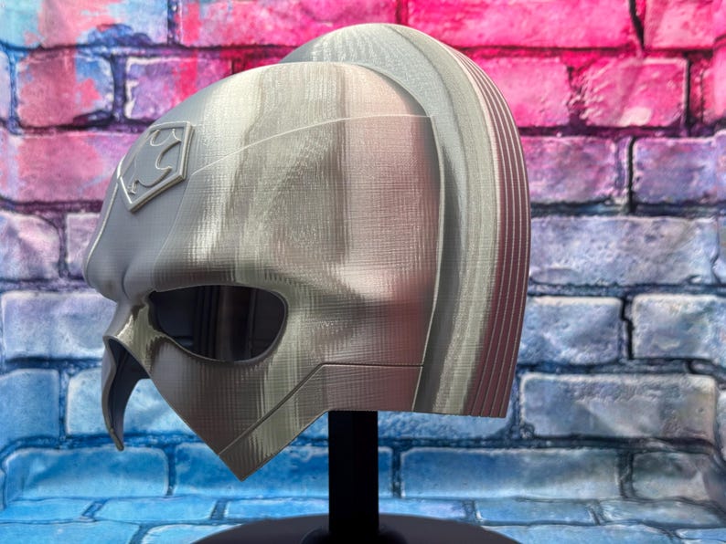 May include: A silver-colored helmet with a Superman logo on the side. The helmet has a smooth, metallic finish and a distinctive design, including eye openings and a pointed chin. The helmet is displayed on a black stand.